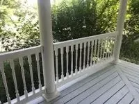 White porch with vertical spindles and support columns; green foliage in background.