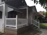 White house with porch and railing, a gray awning and a walkway in front.