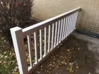 White railing next to a brick building and a bush, with fallen leaves on the ground.