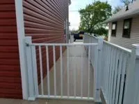 White railing gate along a pathway beside a red-sided building, leading to a house.