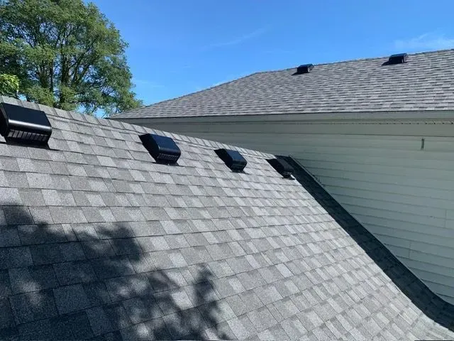 Gray shingled roof with black vents against a blue sky, next to a white house wall.