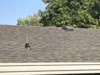 Brown shingle roof with a vent and a small hole, set against a backdrop of trees and a blue sky.