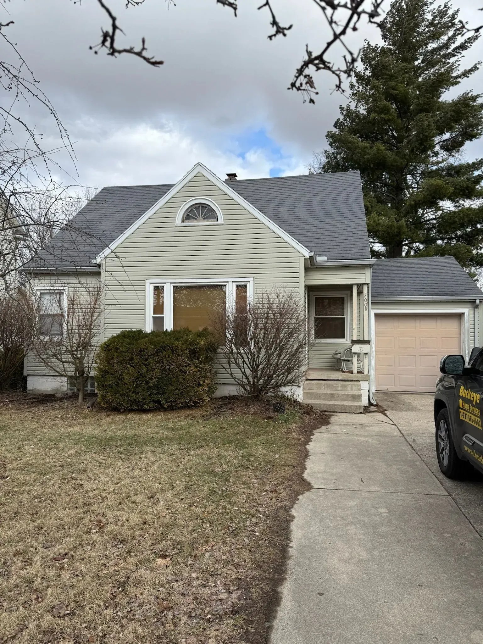 A weathered, single-story house with a garage and a driveway under a cloudy sky.