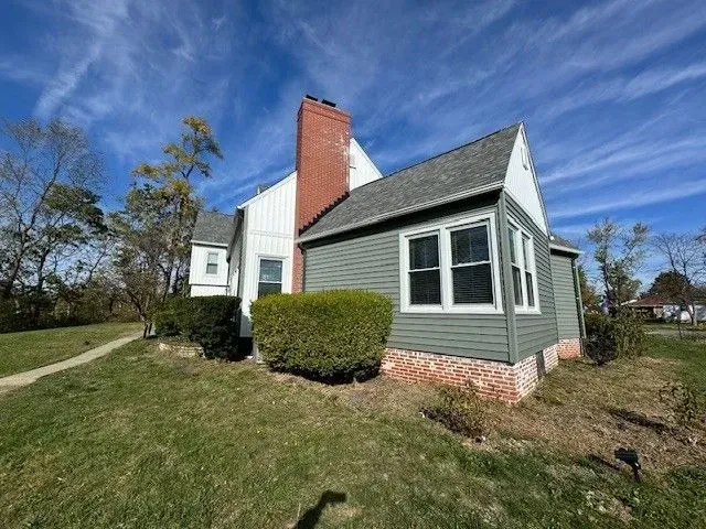 House with green siding and a brick chimney on a grassy lot under a blue sky.