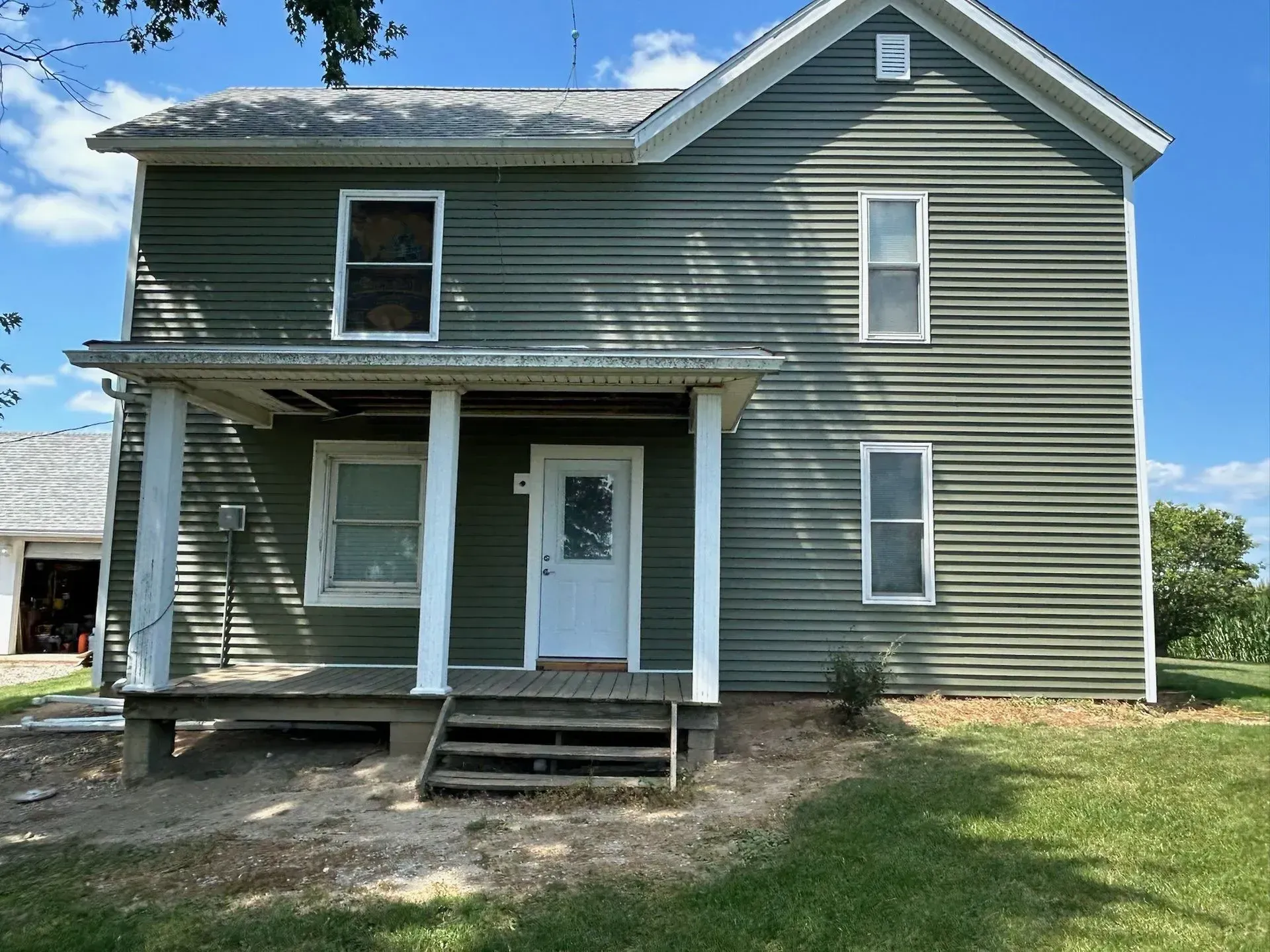 Green two-story house with white trim, porch, and door on a grassy lawn under a blue sky.