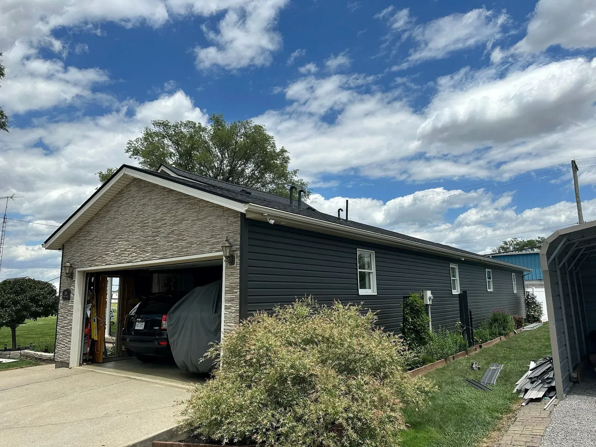 Garage with black siding and stone facade, parked car under a carport, blue sky.
