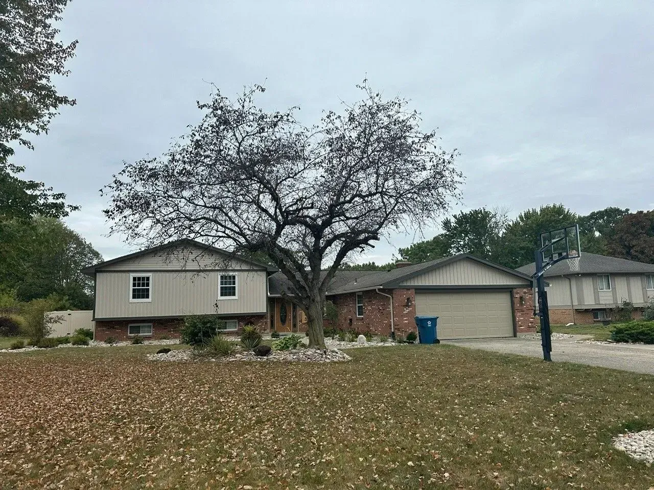 House with beige siding and brick facade, large tree, and driveway on an overcast day.
