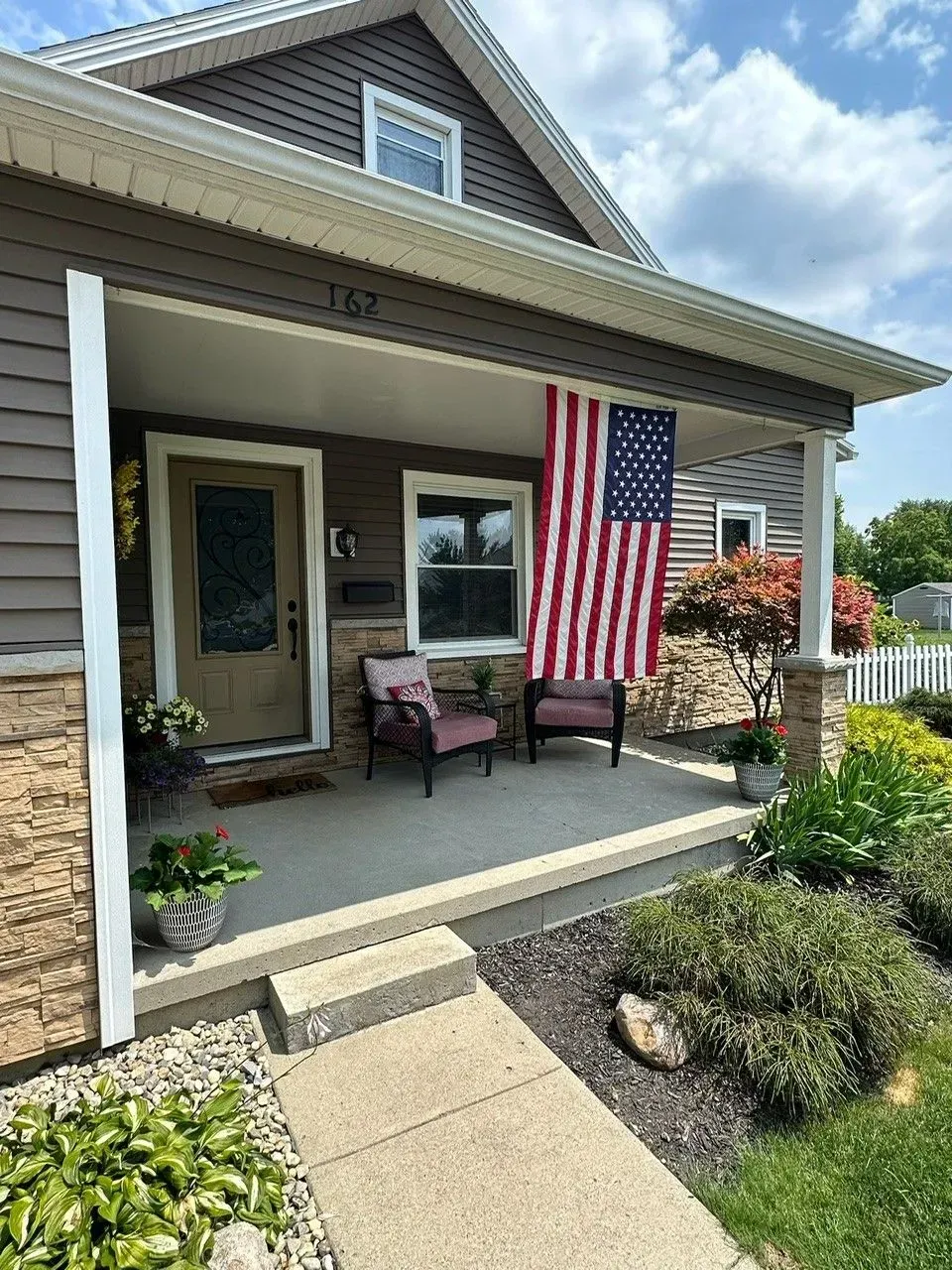 Cozy porch of a home with American flag, two chairs, and landscaping. Brown and beige tones.
