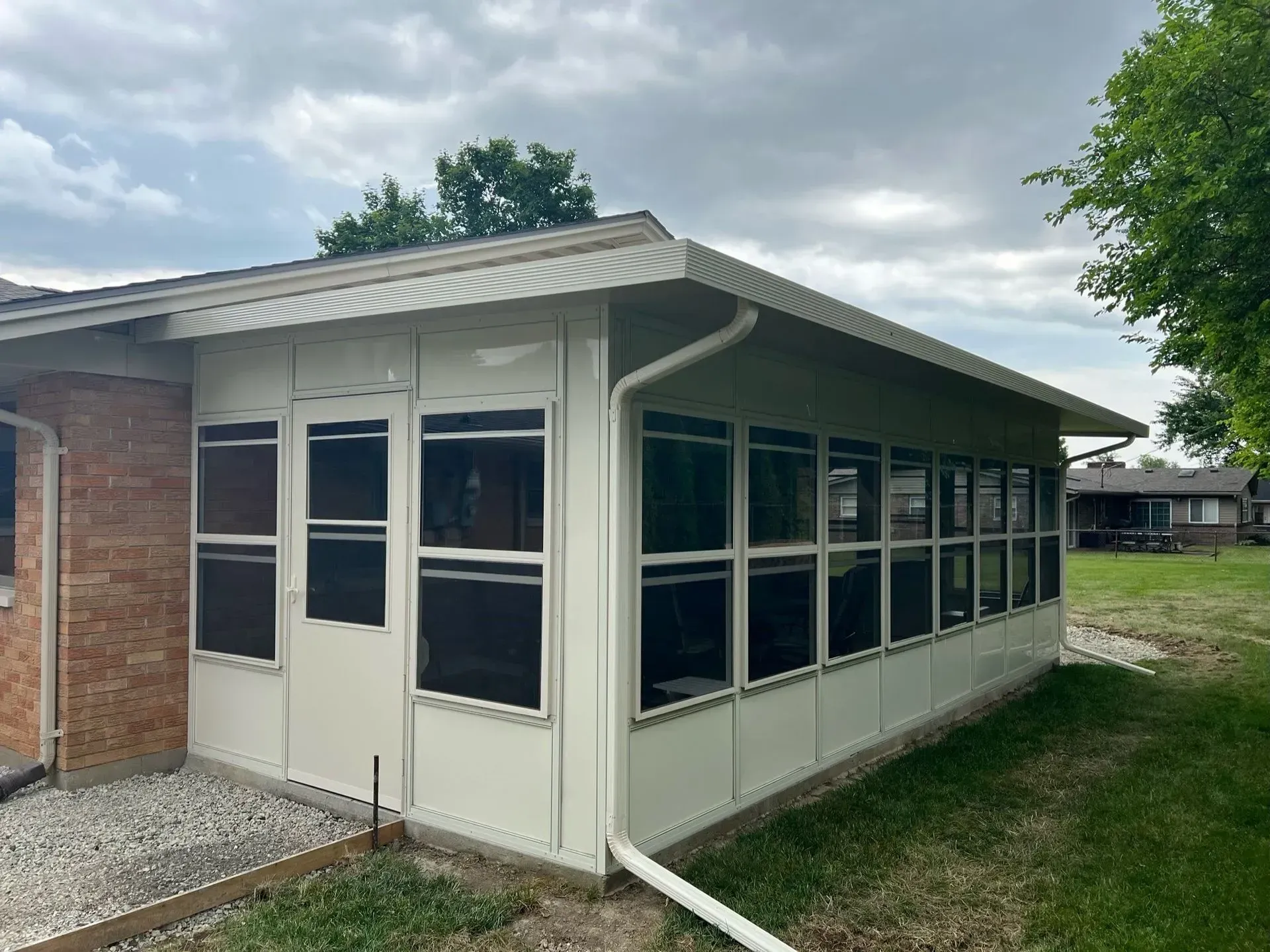 Sunroom addition with many windows, cream siding, and brick wall on a cloudy day.