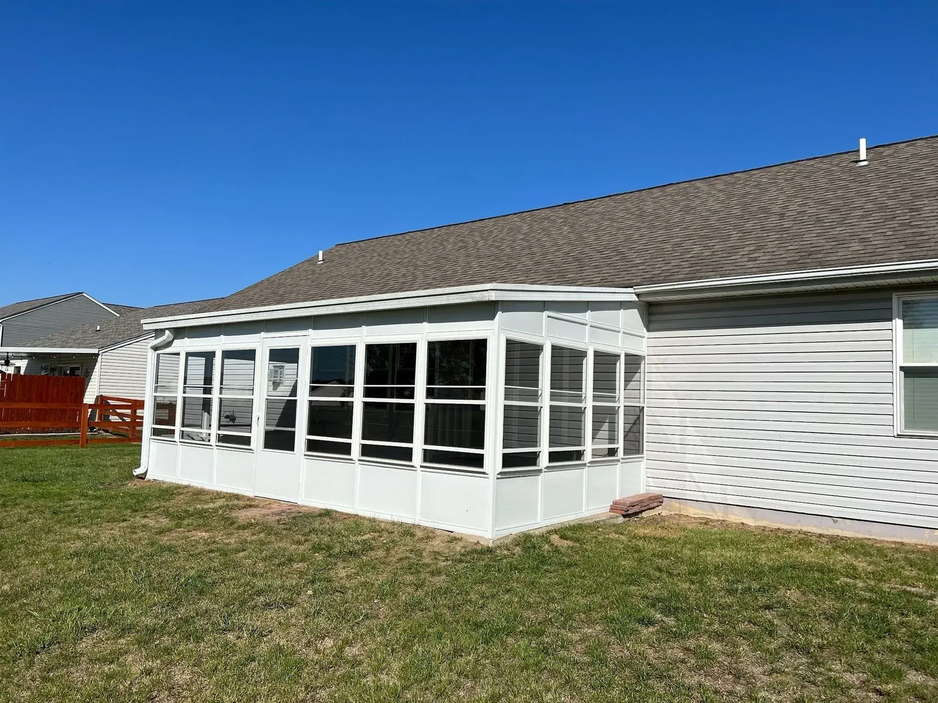 A white sunroom addition attached to a gray house, with windows, on a green lawn. Blue sky.