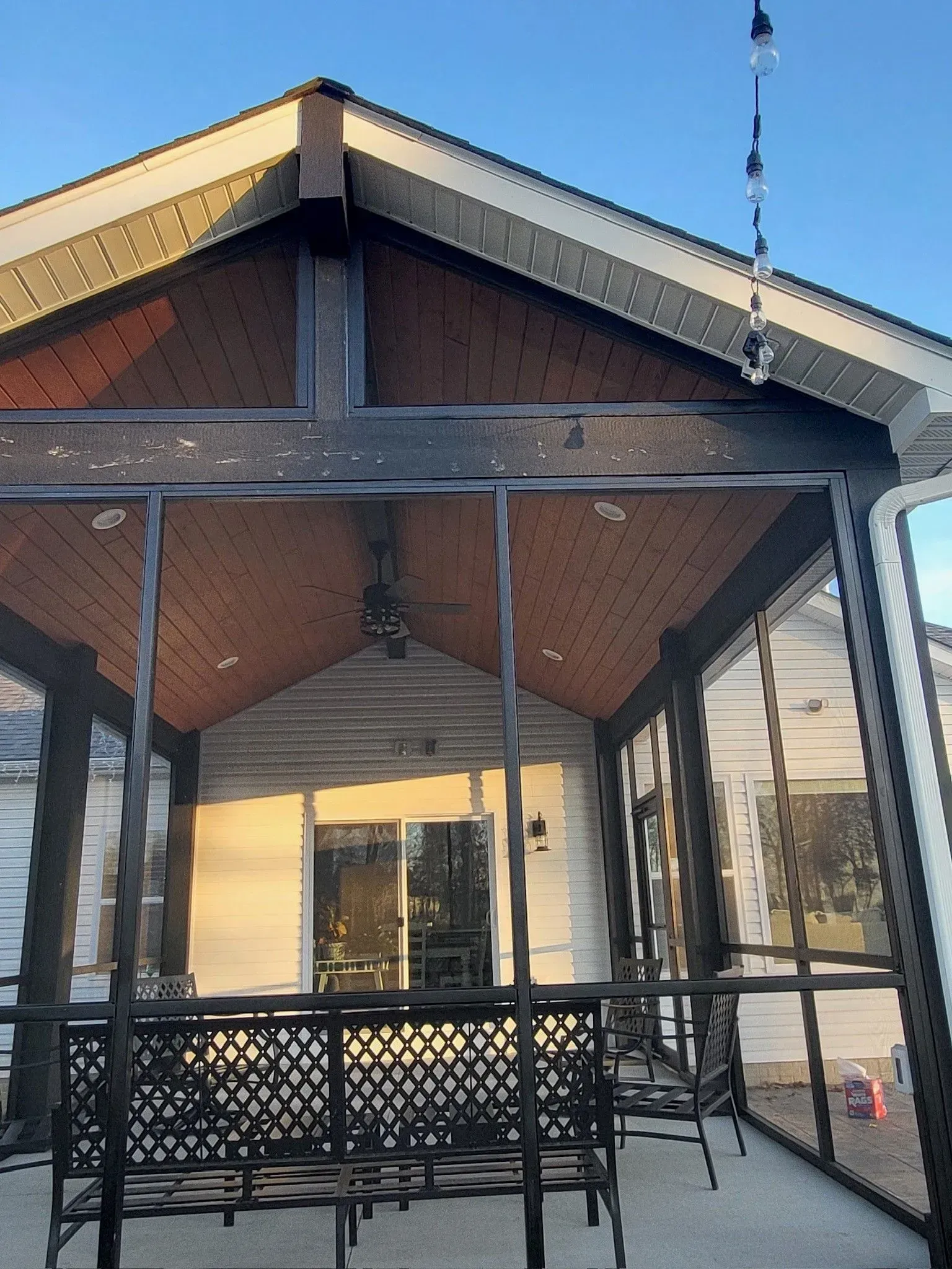 Screened-in porch with brown ceiling, black framing, and a black bench. White house exterior.
