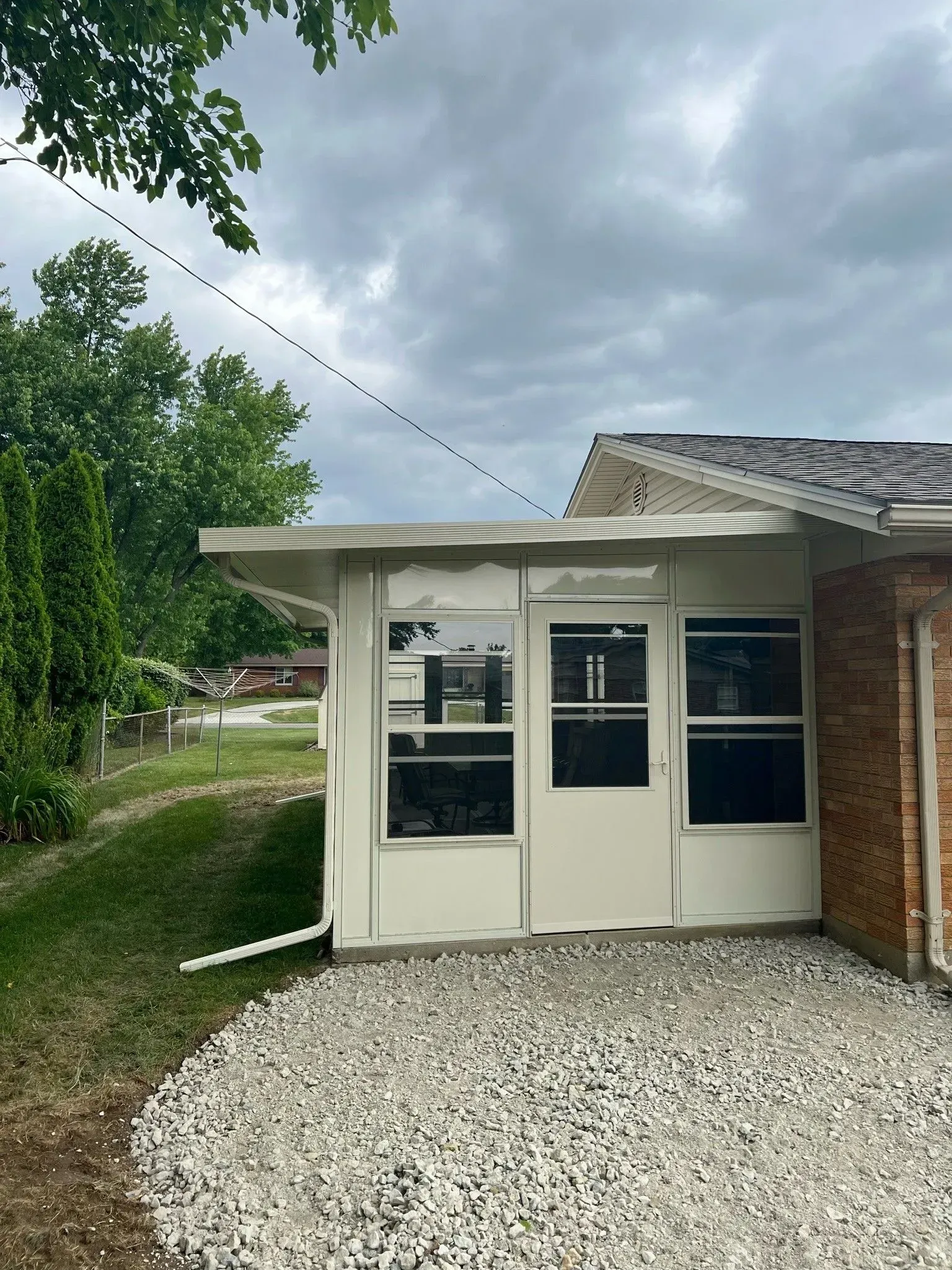 A white sunroom with windows attached to a brick building, gravel ground, overcast sky.