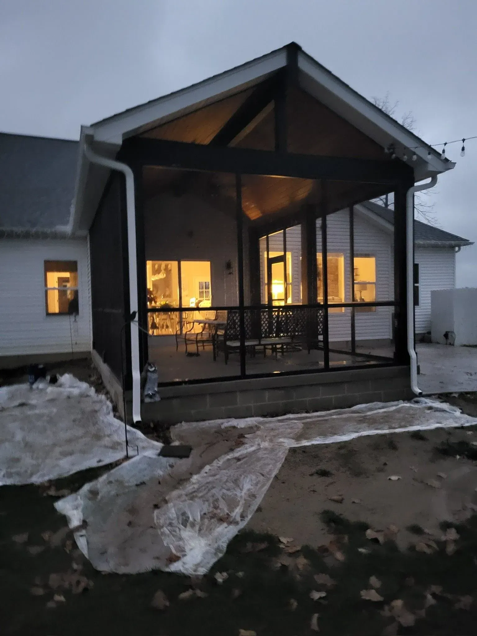 Screened-in porch on a white house, lights on inside. Black frames, brown roof, and melting snow on the ground.