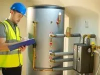 A worker in a hard hat and safety vest inspects a water heater, taking notes on a clipboard in a utility room.