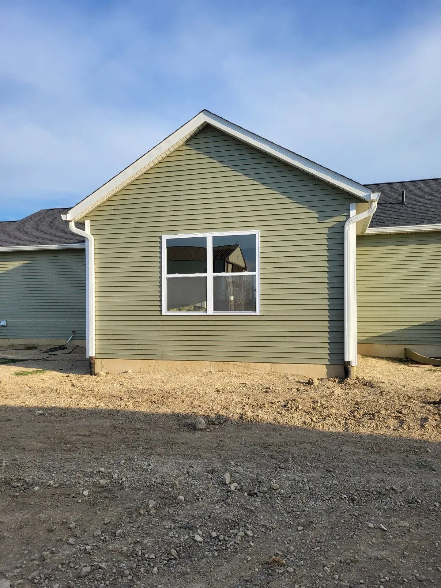 Small building with green siding, white-framed window, and gravel yard under a blue sky.