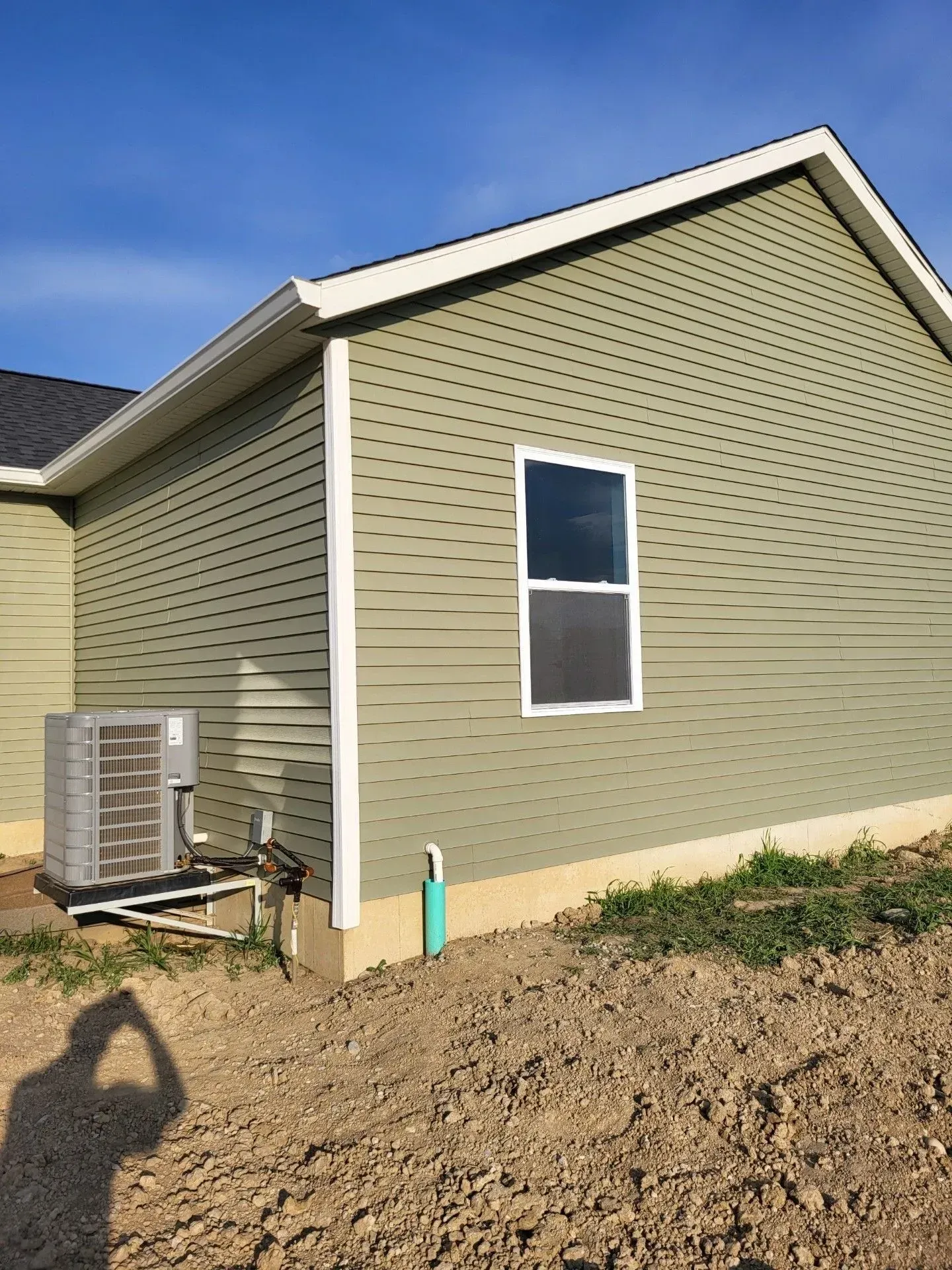 Green-sided house with white trim, window, and air conditioning unit. Dirt mound in front.