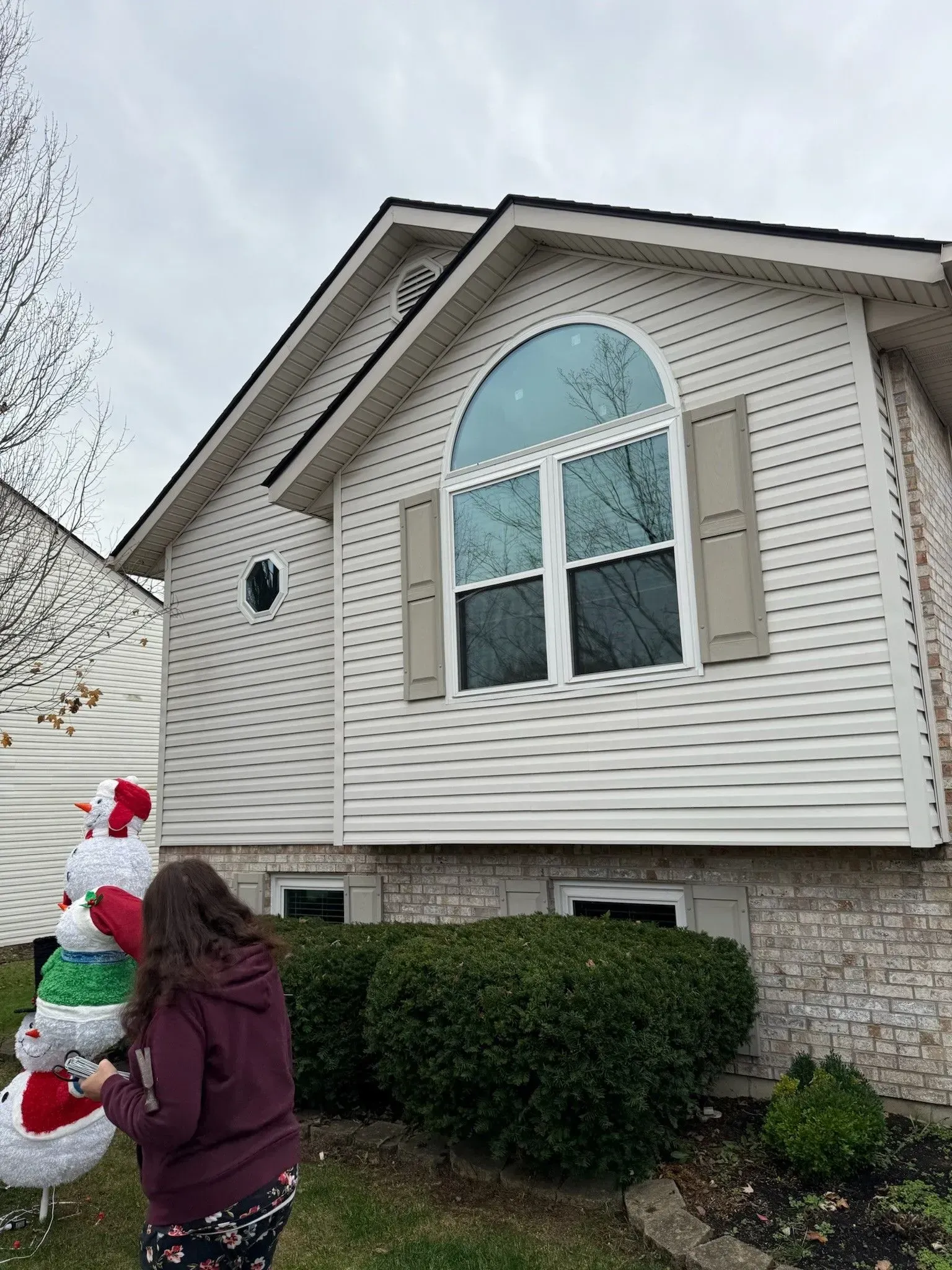 Person near a house with a snowman decoration. The house has a large arched window with shutters. Overcast sky.