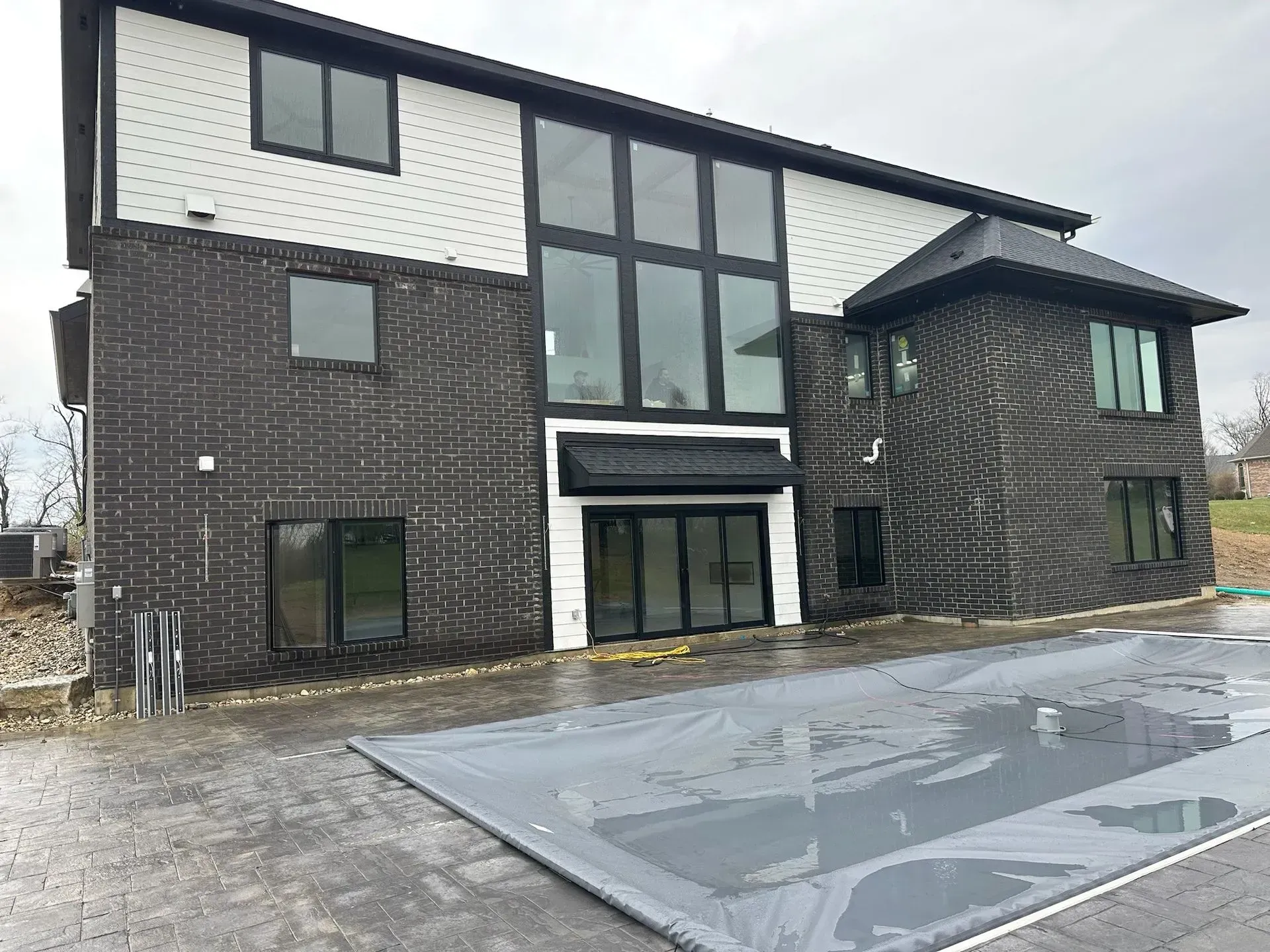 Two-story house exterior with black brick and white siding. Large windows, pool covered. Overcast day.