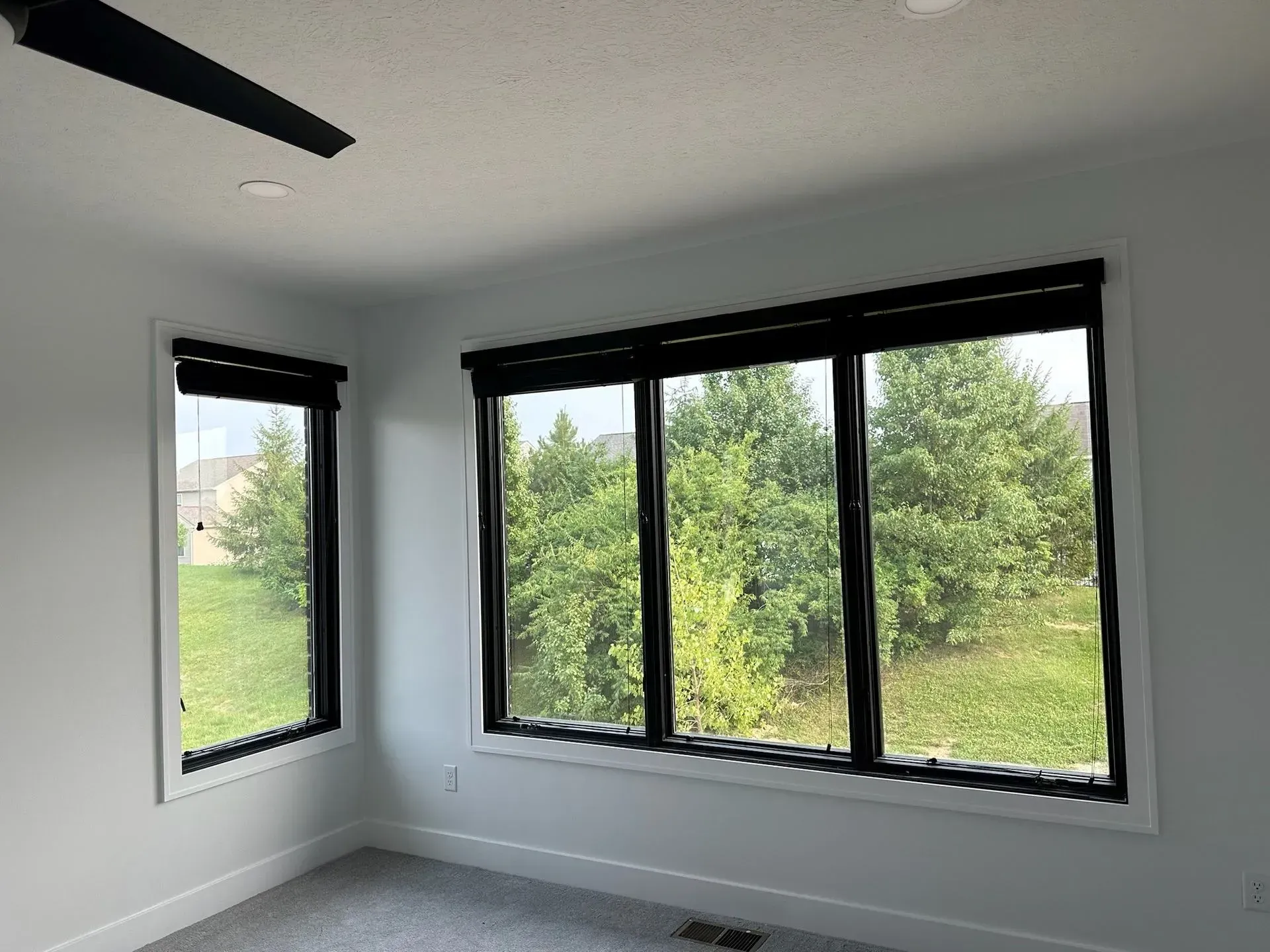 Bedroom with black-framed windows, black shades, white walls, and a view of green trees.