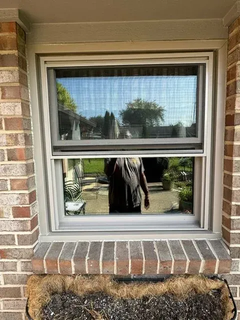 A rectangular window on a brick building with a partially opened screen. The outside view reflects a person and backyard.