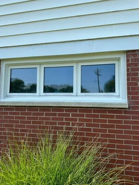 Three-pane white-framed window on a red brick wall, reflecting trees and sky. White siding above, green grass below.