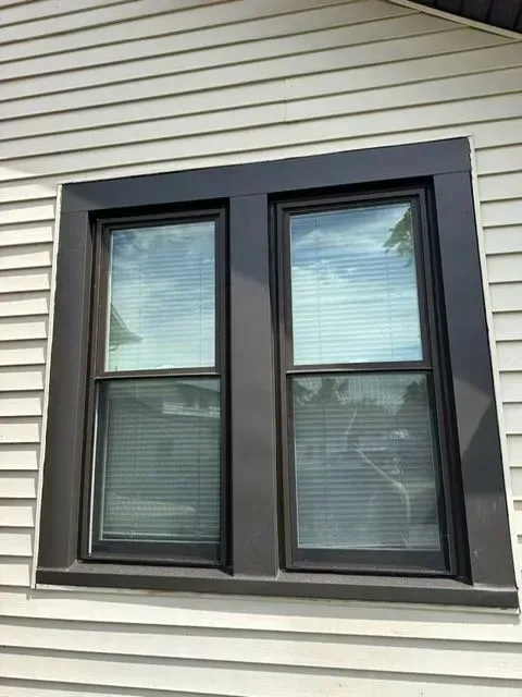 Two black-framed windows on a white-sided house. Reflective glass shows blinds and sky.