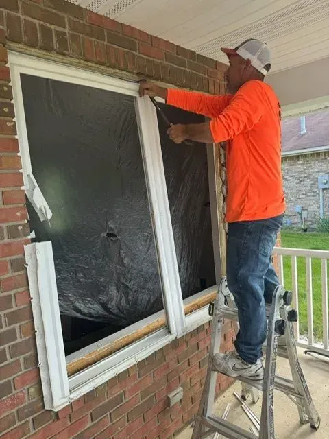 Person on ladder installing window, exterior setting, orange shirt, brick wall, white trim.