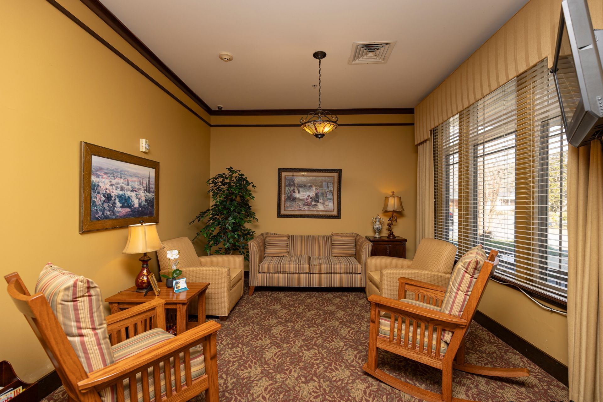 A hotel lobby seating area with a sofa, two wooden rocking chairs, a side table, and wall art in a room with tan walls.