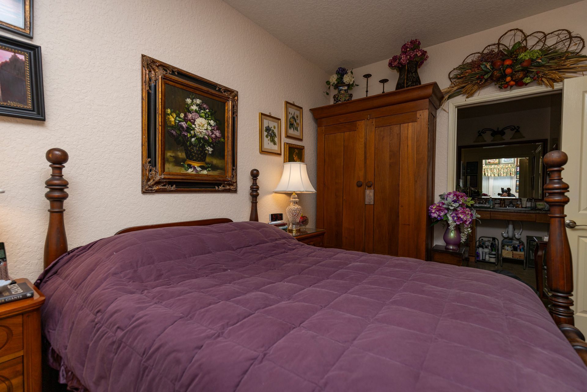A bedroom with a wooden four-poster bed covered by a purple quilt, a wooden wardrobe, and framed art on the walls.