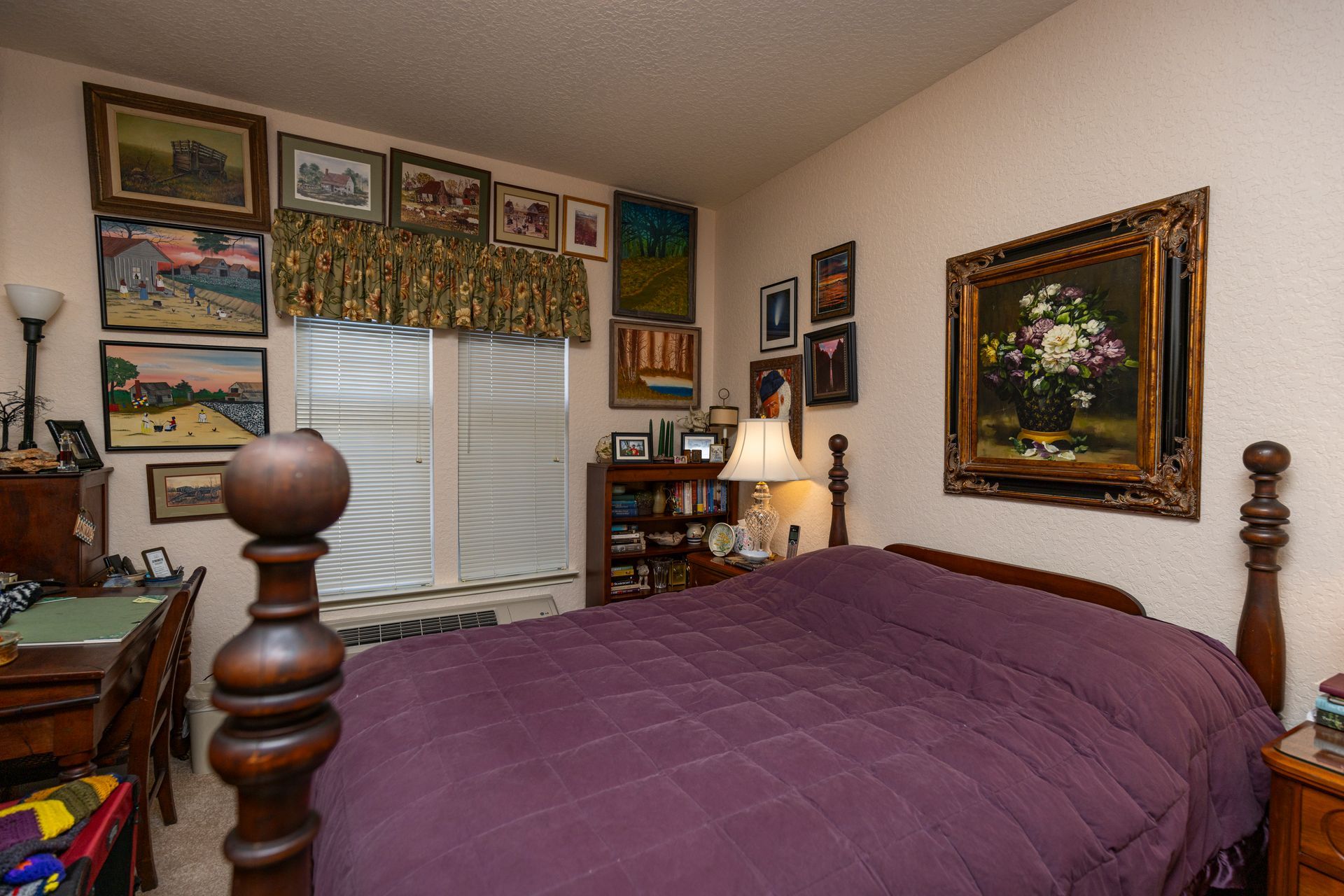A bedroom featuring a bed with a purple quilt, a wooden headboard, a corner desk, and many framed art pieces on the walls.