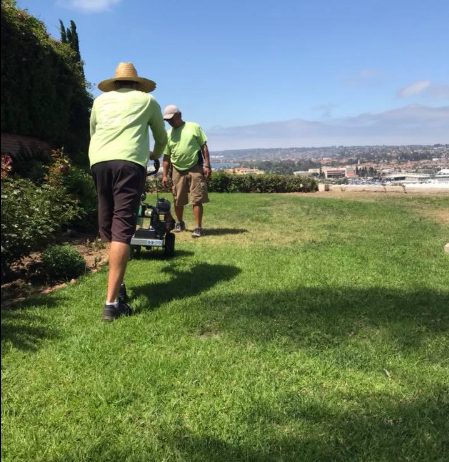 Two people mowing grass on a sunny hillside overlooking a city.