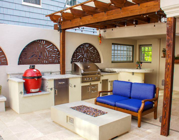 Outdoor kitchen with grill, red smoker, and seating area with blue cushions. Wooden pergola overhead.