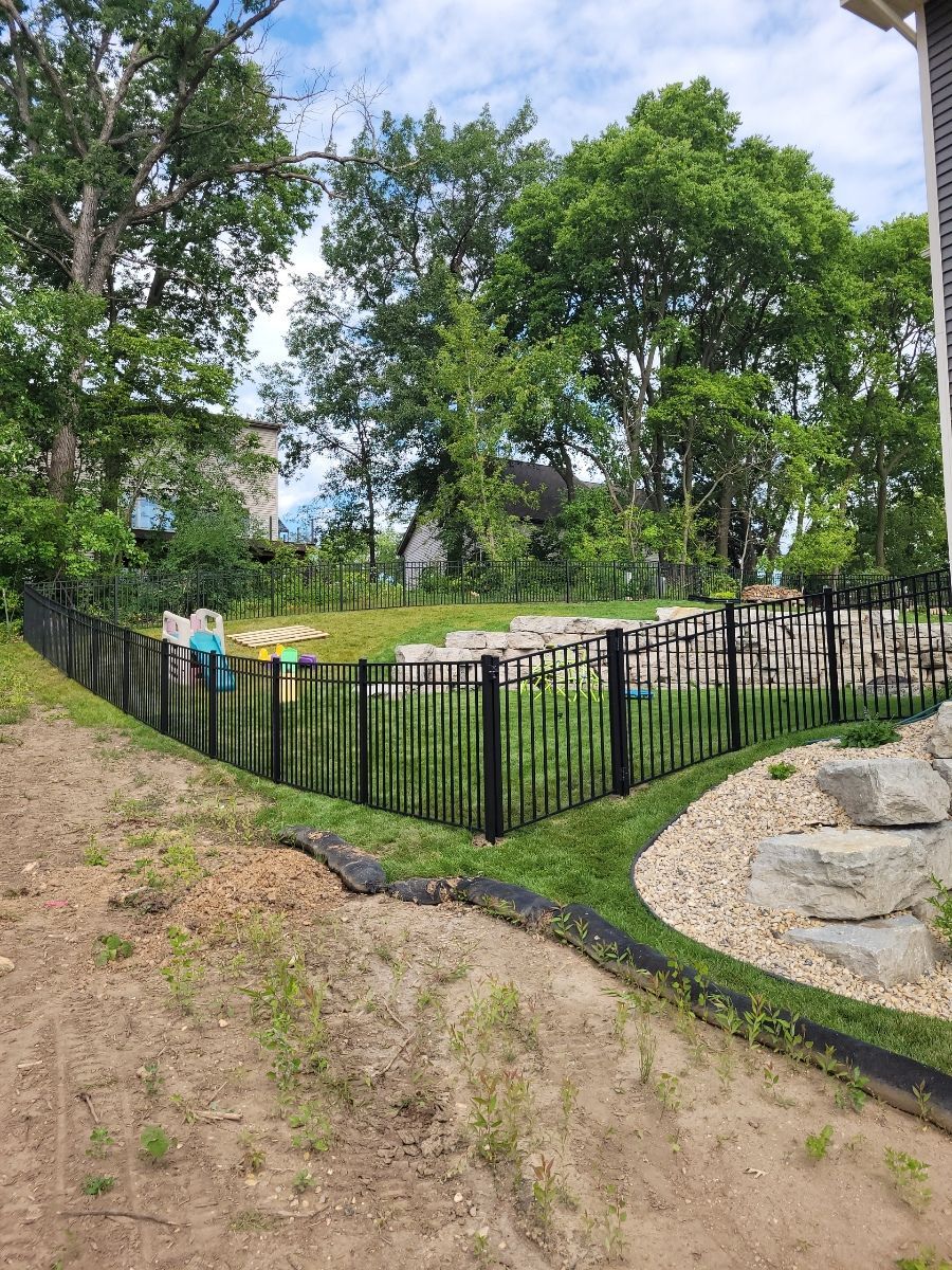 A black fence surrounds a backyard with trees and rocks.