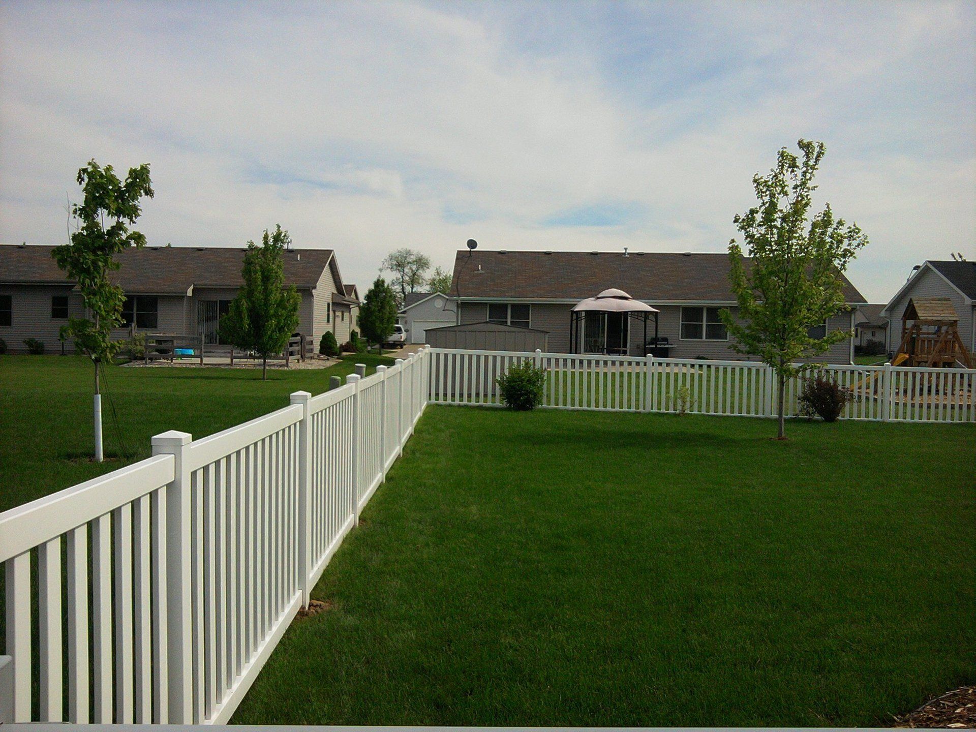 A white fence surrounds a lush green yard with houses in the background