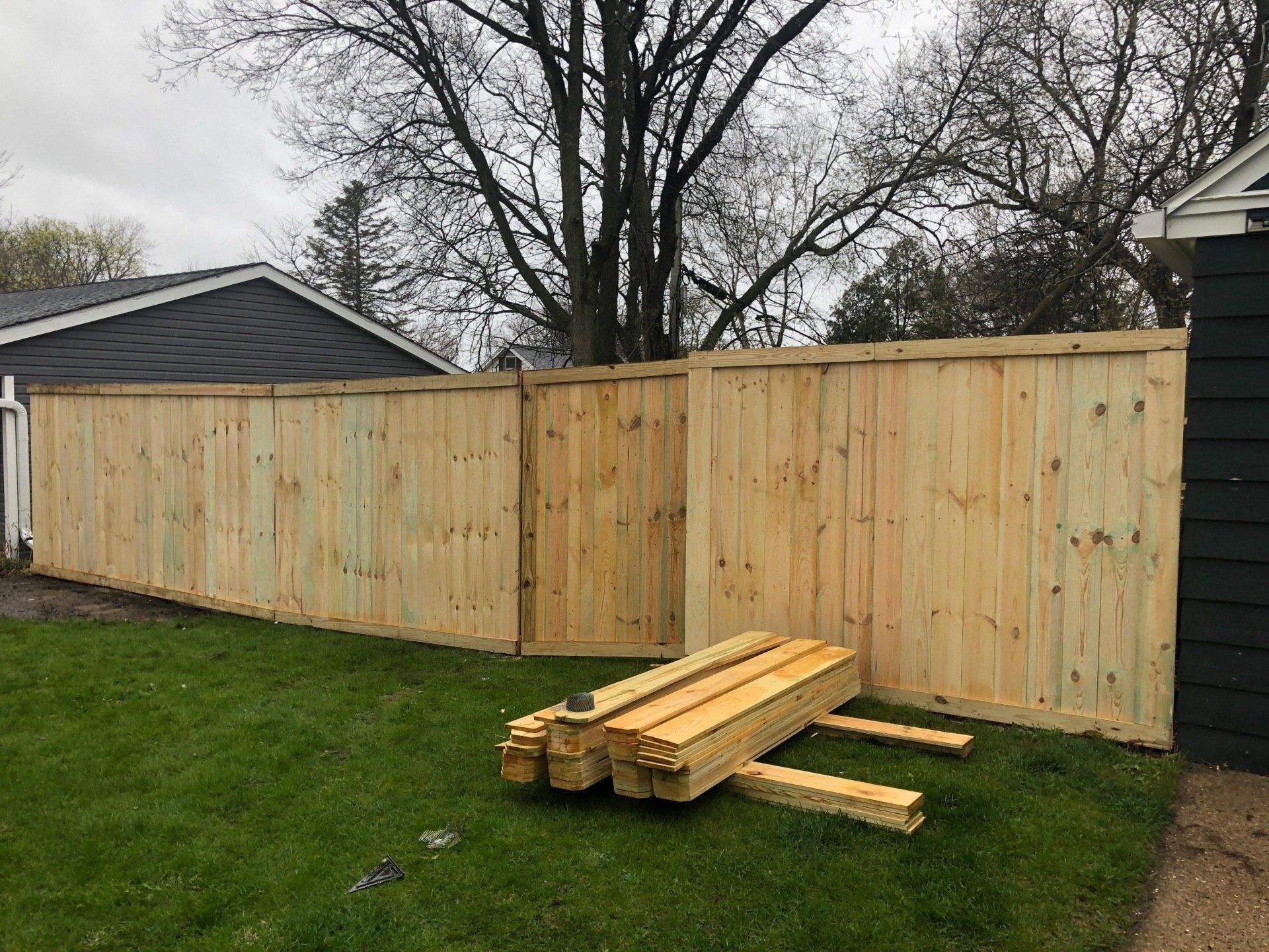 A wooden fence is being built in a backyard with a pile of wood in front of it.