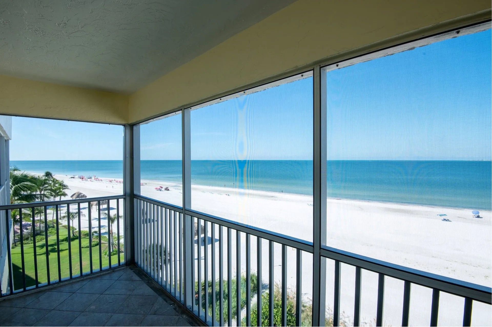 A screened-in balcony overlooking a sunny, white sand beach and the ocean.