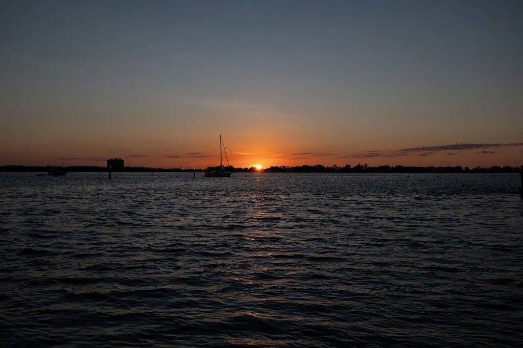 A sunset glows over a dark, rippling body of water, with the silhouettes of boats visible on the horizon.