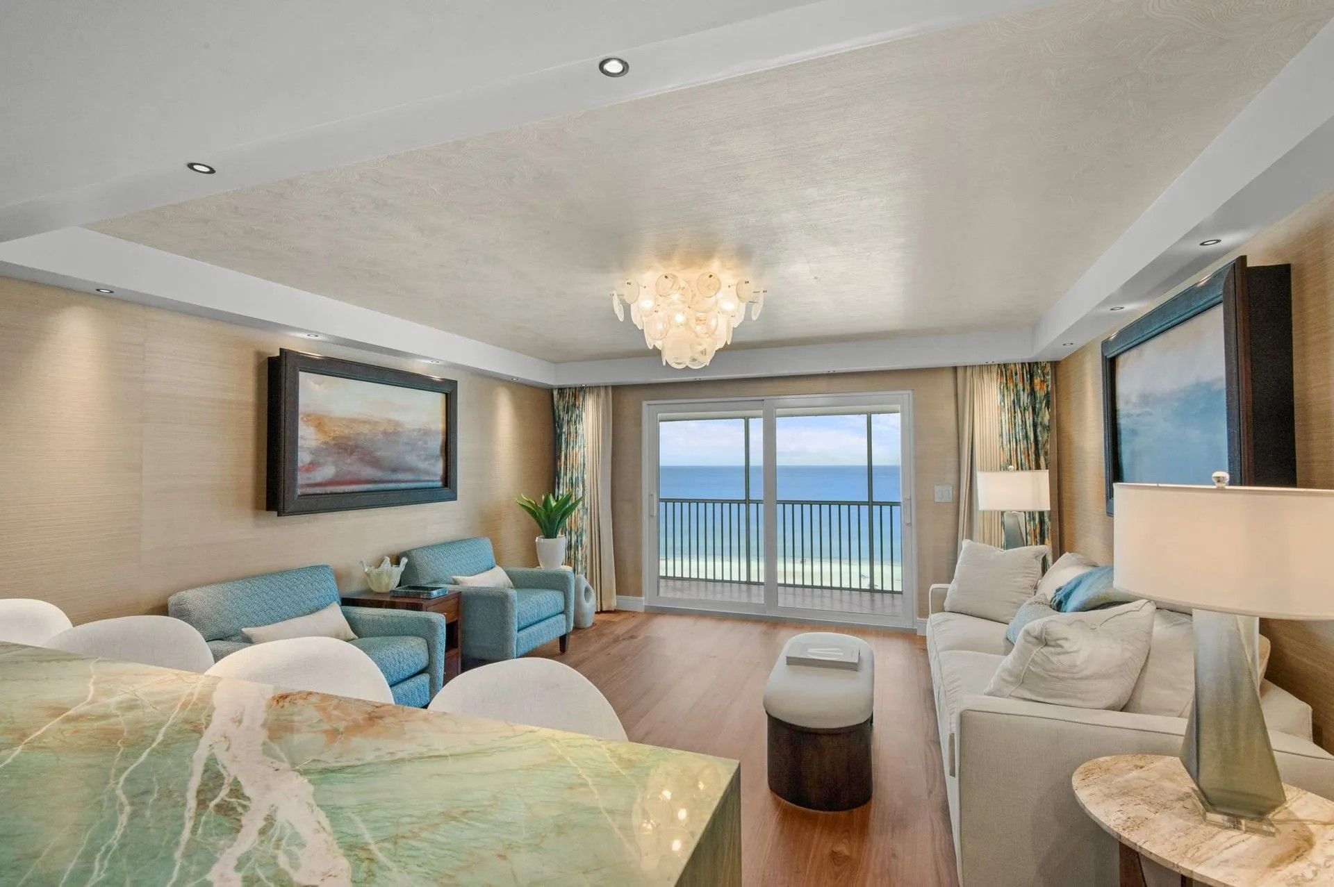A beach-view hotel living room with light-toned furniture, turquoise accents, a chandelier, and a sliding glass door.