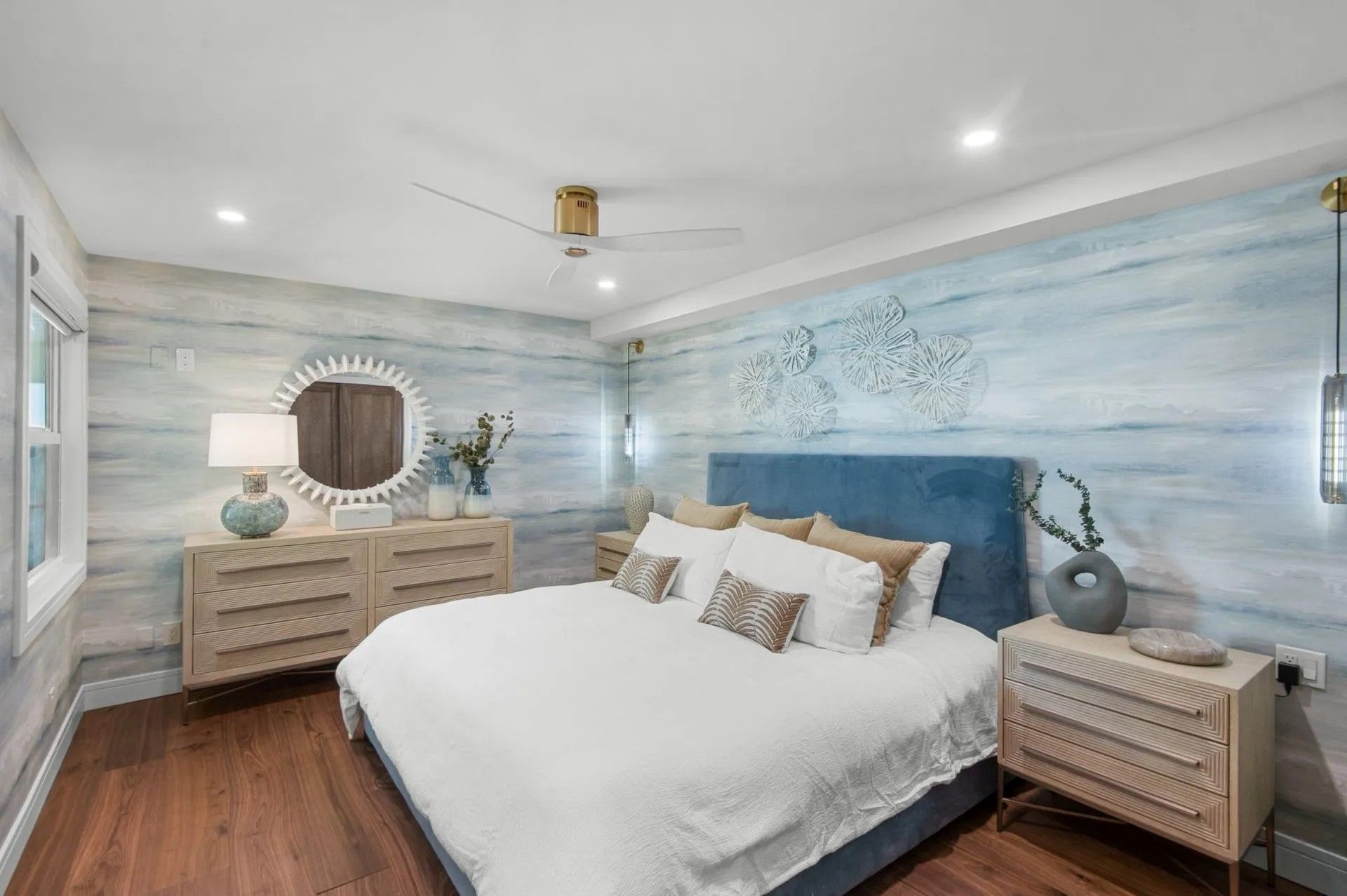 A bedroom featuring a blue upholstered bed, light wood nightstands, and walls with blue and white watercolor-style paint.