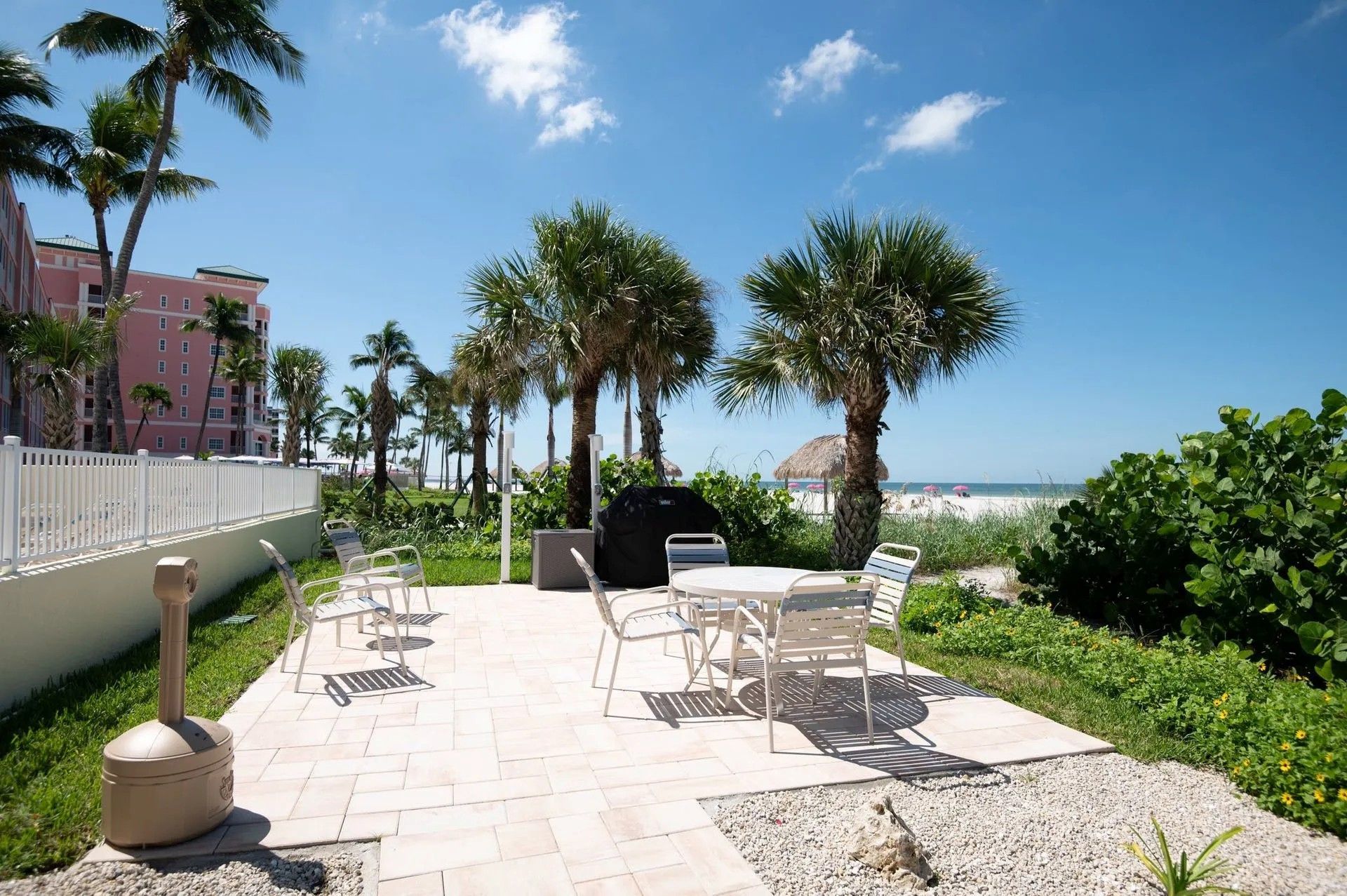 A sunny outdoor patio with chairs and a table, featuring palm trees, greenery, and a view of a beach under a blue sky.