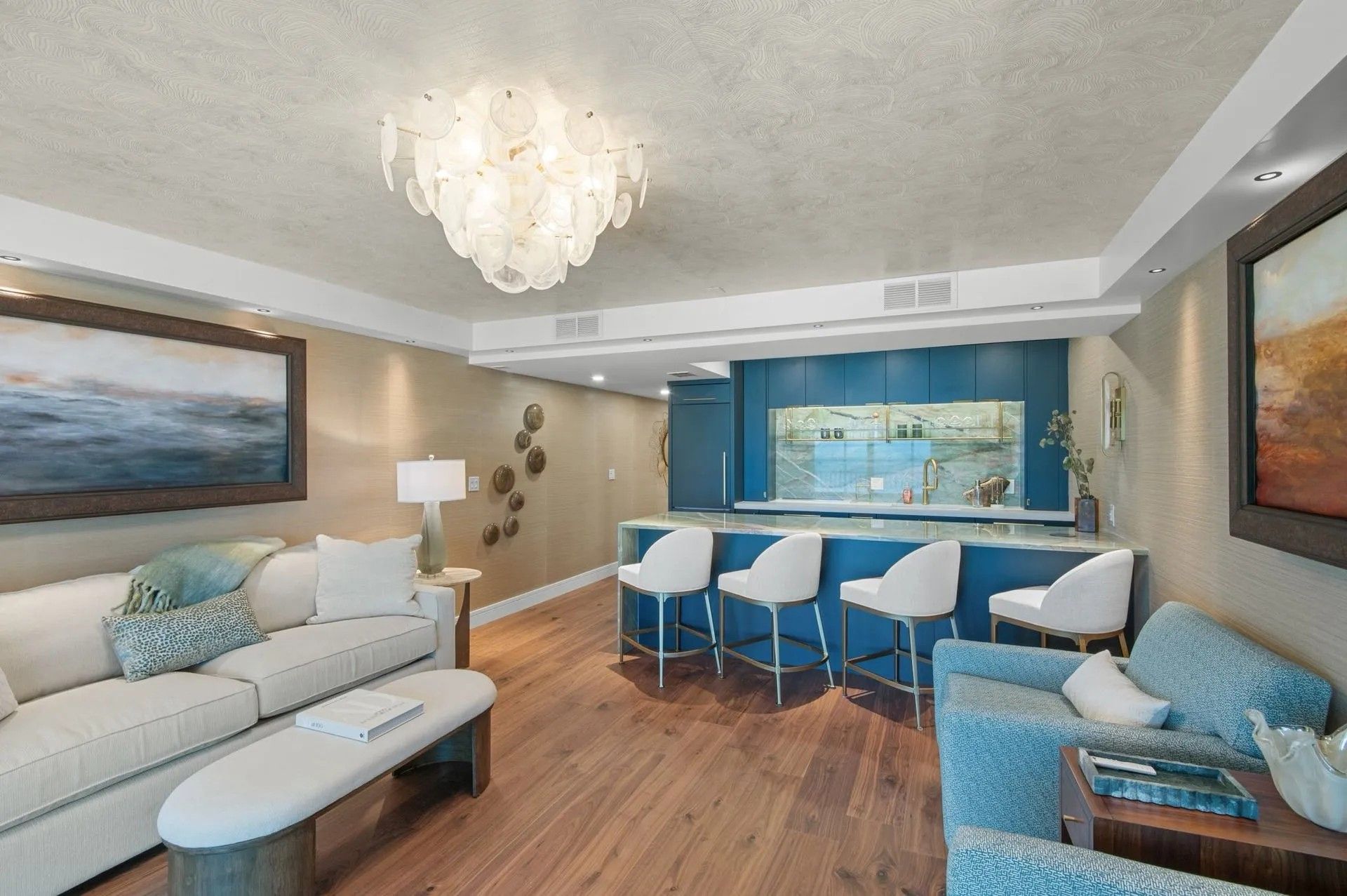 A living area with a sofa, an ottoman, a bar with four white stools, wood flooring, and blue decorative accents.