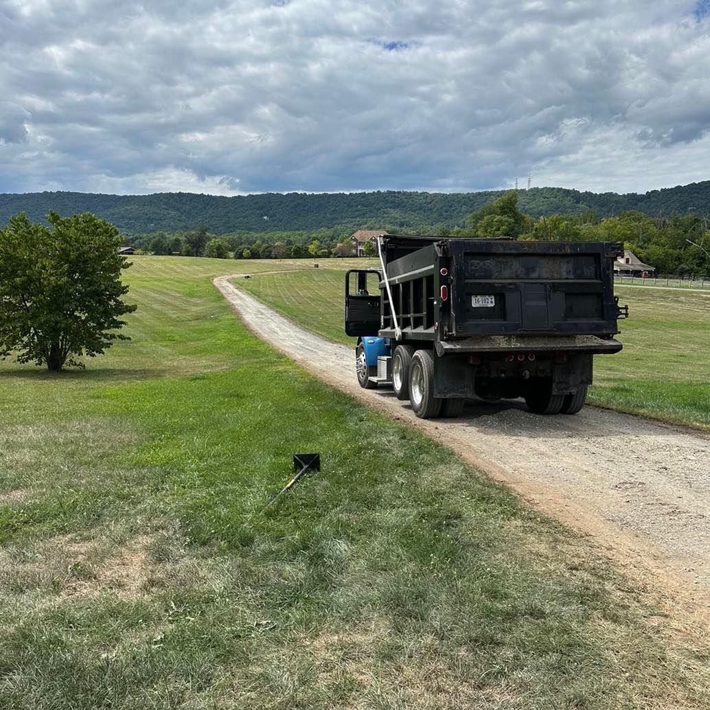 A dark dump truck drives away on a gravel road through a grassy field toward distant mountains under a cloudy sky.