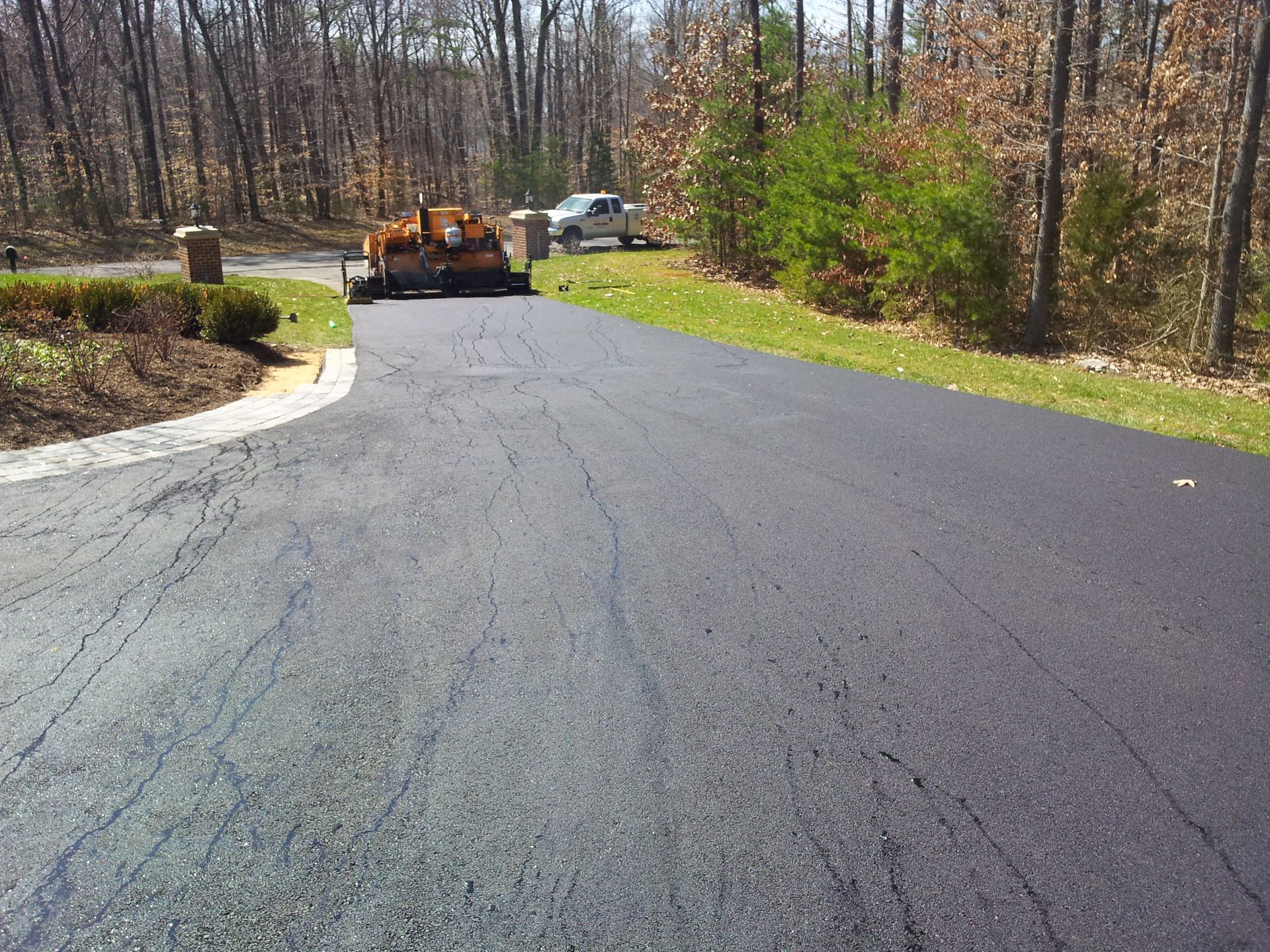 A long, paved asphalt driveway leads toward a truck and trees in a sunny, wooded outdoor setting.