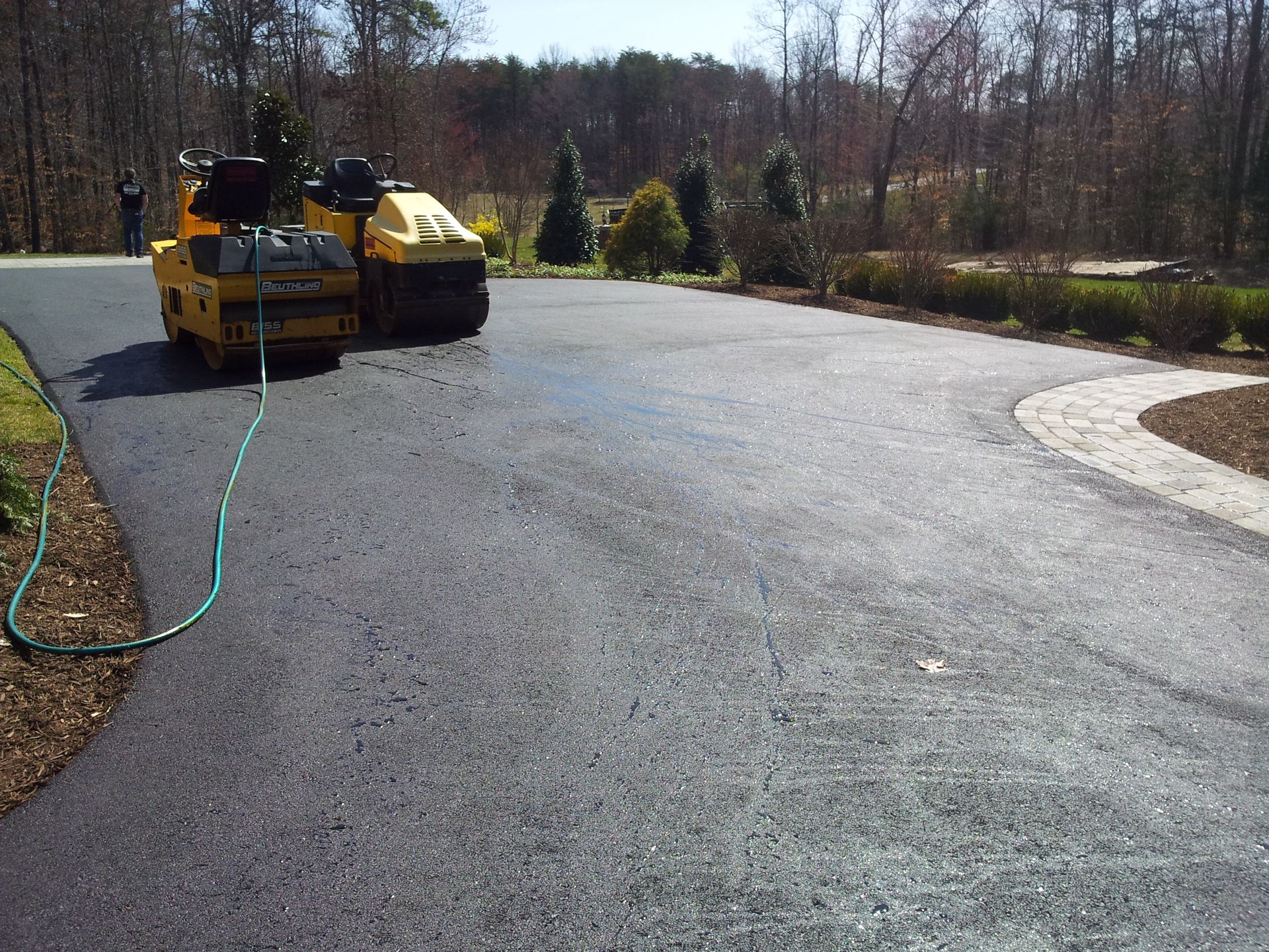Yellow construction machinery on a freshly paved black asphalt driveway near a treeline.