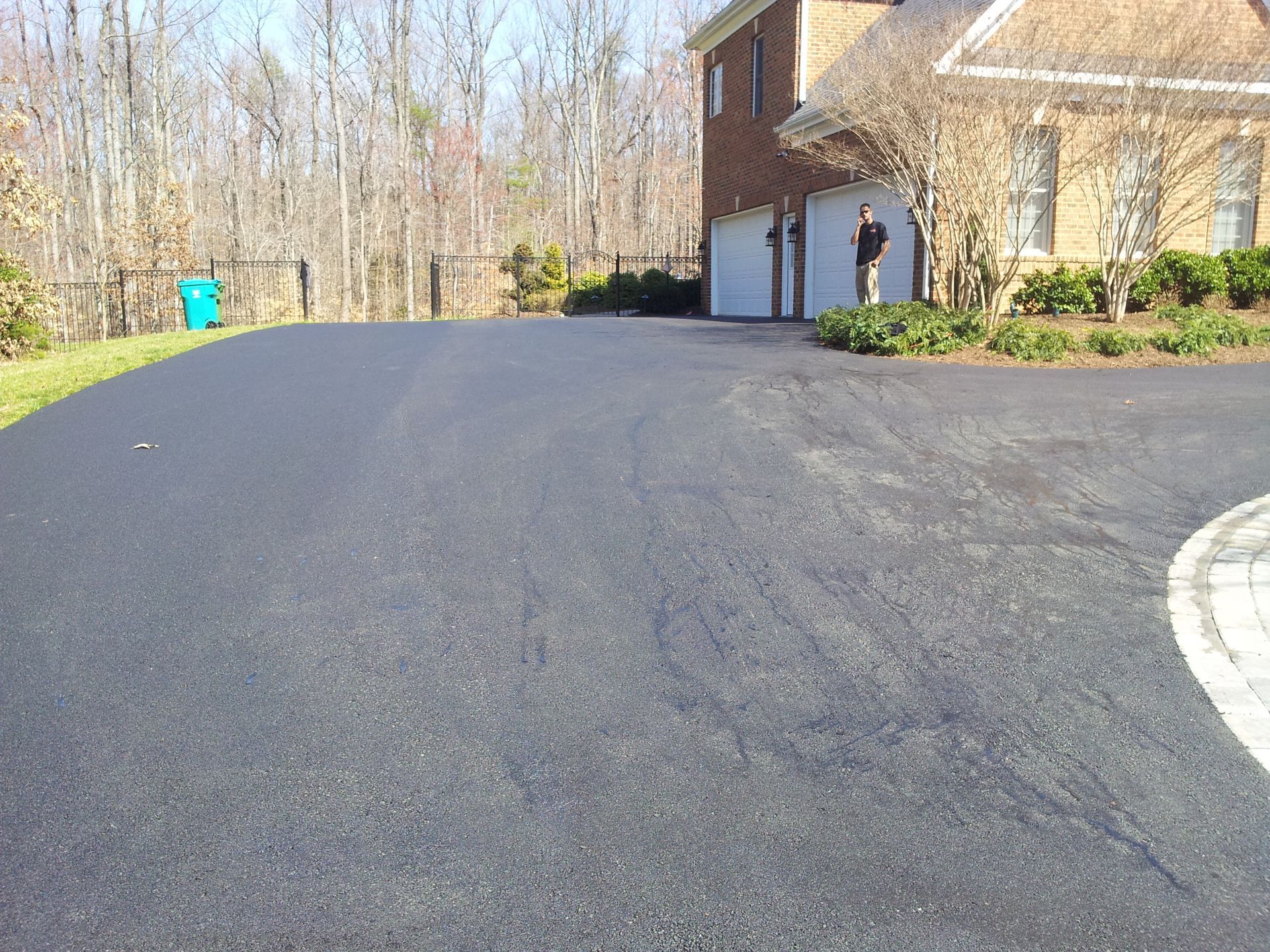 A person stands in a driveway in front of a brick house with a multi-car garage, bordered by trees and a lawn.