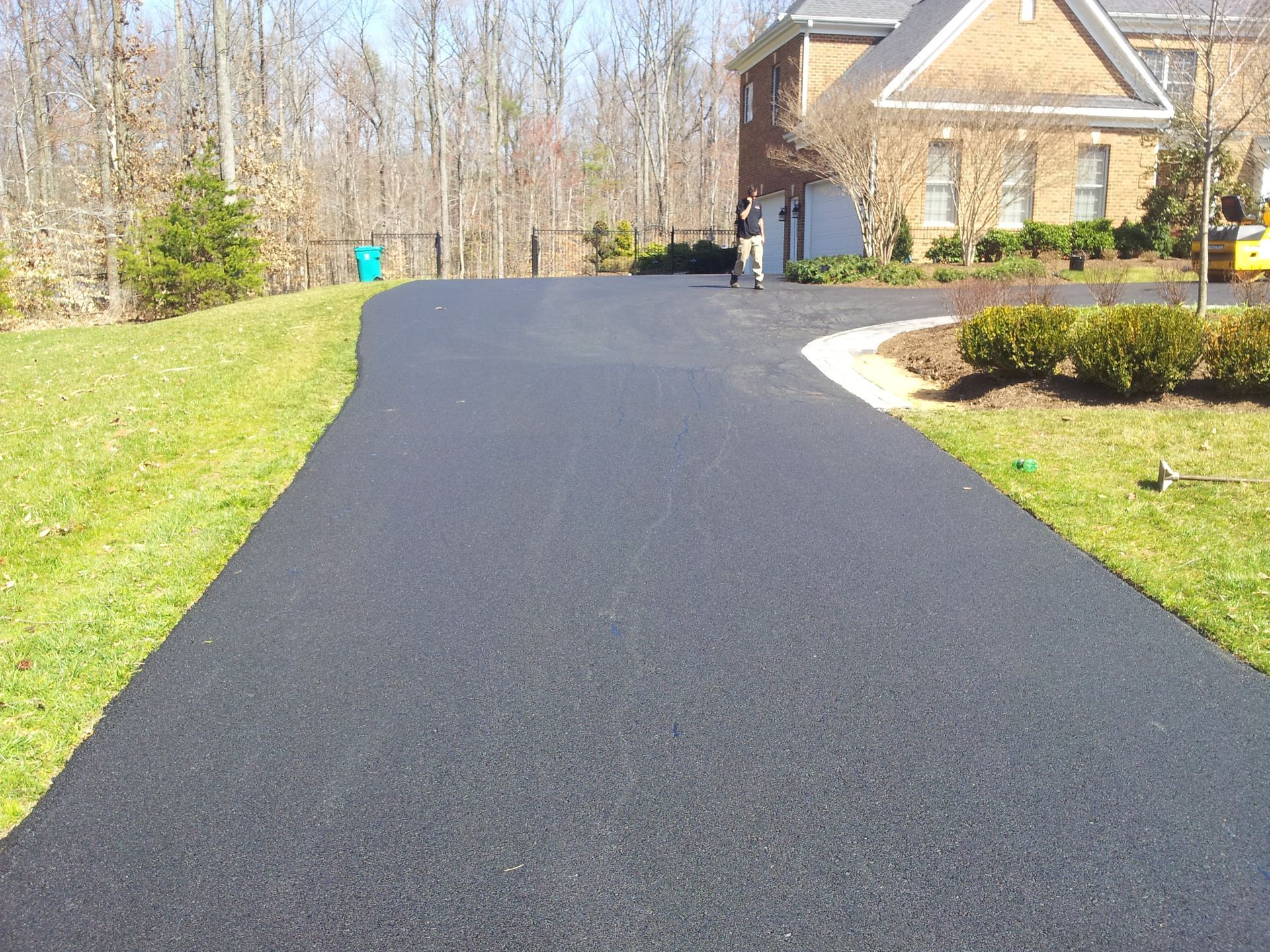 A newly paved dark asphalt driveway leads to a brick house on a sunny day with green lawns on either side.