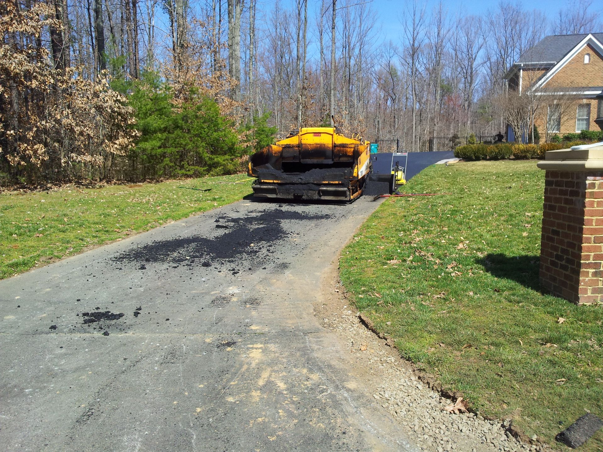 A yellow asphalt paver sits on a residential driveway near a brick pillar, with fresh black asphalt applied in front of it.