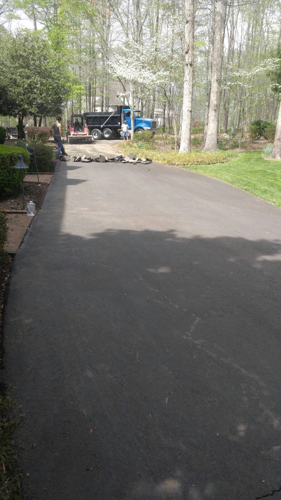A freshly paved asphalt driveway leads toward a blue dump truck parked in a wooded residential area.