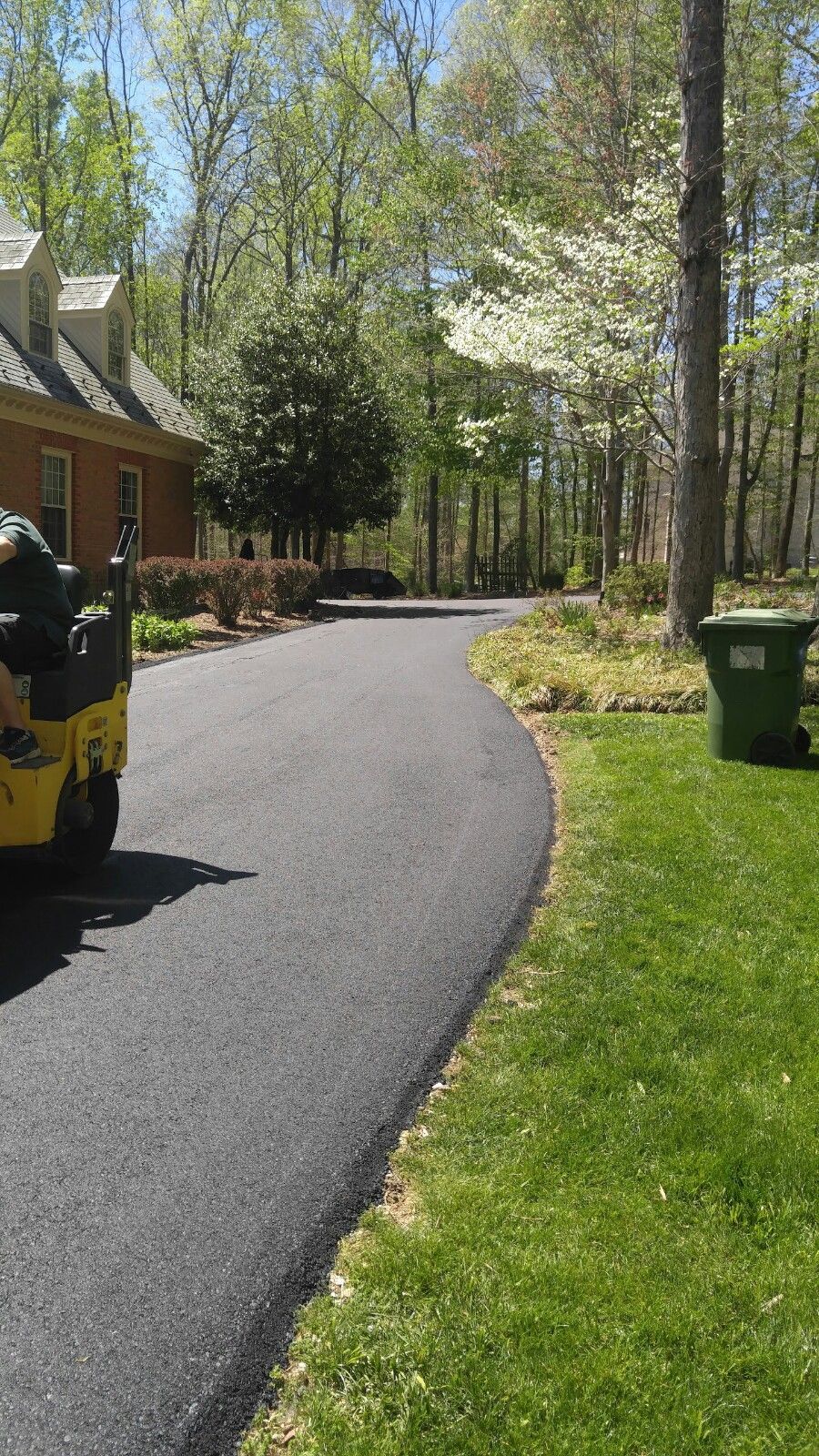 A yellow road roller sits on a freshly paved black asphalt driveway leading to a brick house among trees.