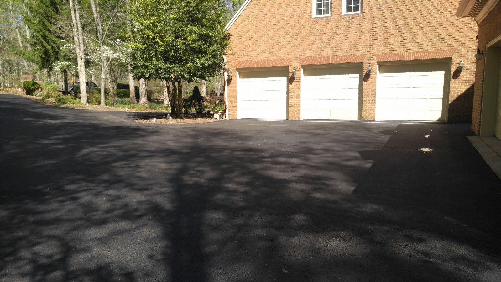 A large, paved driveway leads to a brick house with three closed white garage doors, surrounded by trees.